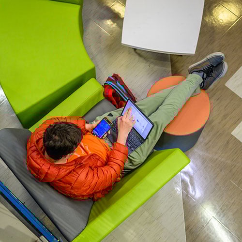 Male student using laptop and mobile device sitting in chair